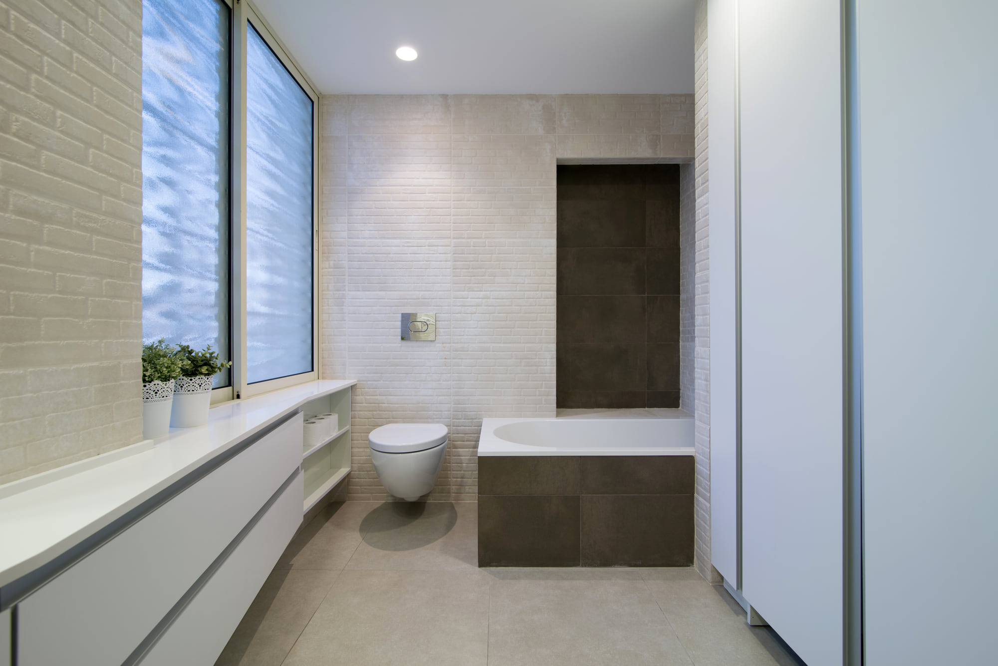The children’s bathroom along its length — a continuous counter beneath the window, a laundry cupboard and a bath against a dark-clad niche