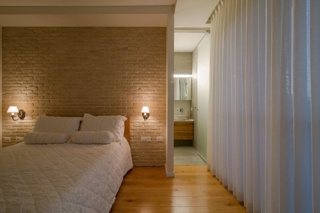 The master bedroom — the reclaimed brick wall in concealed light, and on the right the way through to the bathroom behind a linen curtain