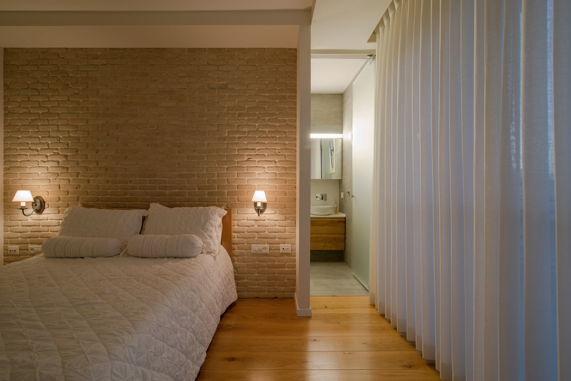 The master bedroom — the reclaimed brick wall in concealed light, and on the right the way through to the bathroom behind a linen curtain